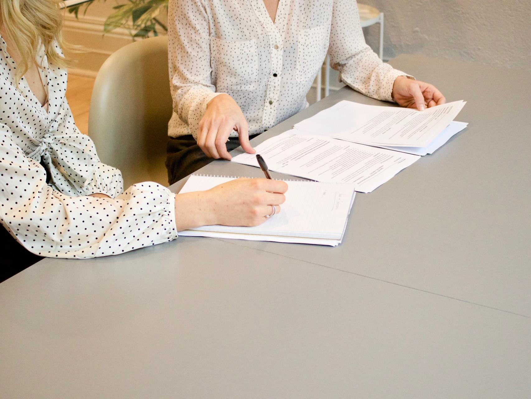 woman signing on white printer paper beside woman about to touch the documents